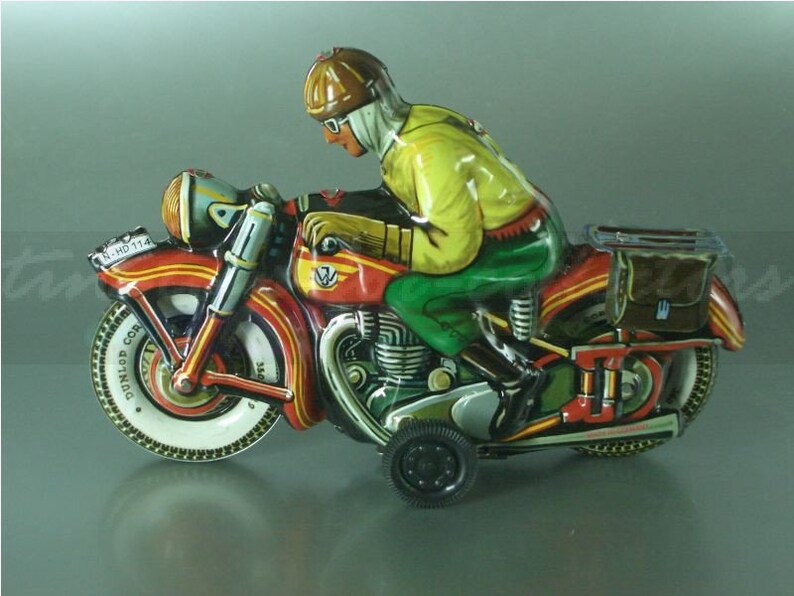 K&ouml;nnte beinhalten: Ein rotes, gelbes und gr&uuml;nes Spielzeugmotorrad mit einem Fahrer, der einen braunen Helm und eine Schutzbrille tr&auml;gt. Das Motorrad hat einen schwarzen Beiwagen mit einer braunen Tasche im Inneren. Das Motorrad hat einen schwarzen Dunlop-Reifen am Vorderrad und einen schwarzen Reifen am Hinterrad. Das Motorrad hat ein schwarzes Nummernschild mit der Aufschrift "N-HO 114".