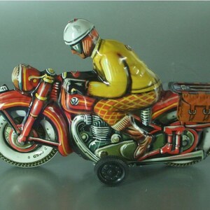 K&ouml;nnte beinhalten: Ein rotes und schwarzes Spielzeugmotorrad mit einem Fahrer, der einen gelben und wei&szlig;en Helm tr&auml;gt. Das Motorrad hat eine braune Satteltasche und einen wei&szlig;en Reifen mit schwarzer Beschriftung "340 x 120".