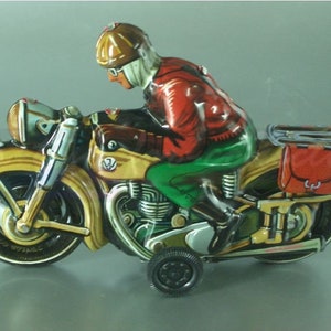 K&ouml;nnte beinhalten: Ein vintage Blechspielzeug-Motorrad mit einem Fahrer, der einen braunen Helm und eine rote Jacke tr&auml;gt. Das Motorrad ist gelb und gr&uuml;n mit einer roten und braunen Satteltasche.