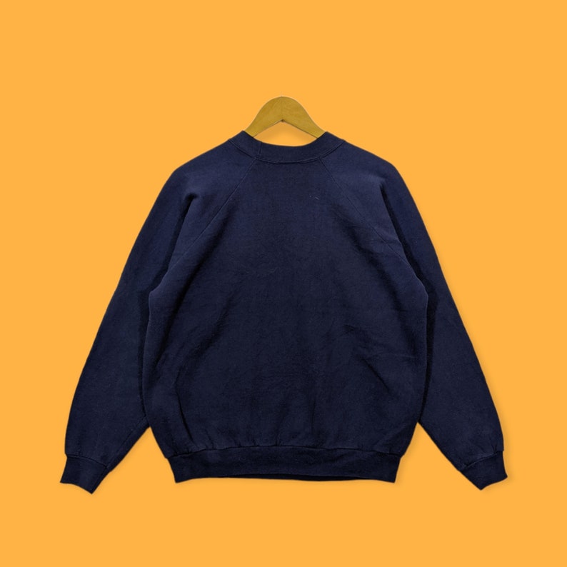 May include: A navy blue sweatshirt with long sleeves, displayed on a wooden hanger against an orange background. The sweatshirt is made of a soft material and has a crew neck.