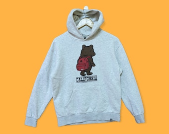 Peut inclure: Un sweat à capuche gris avec un ours brun portant un sac à dos rouge et le mot "CALIFORNIA" imprimé sur le devant.