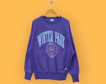 May include: Purple sweatshirt with the text "WINTER PARK" in white outlined in blue. A crest with a mountain range and the text "WINTER PARK" is below the text.