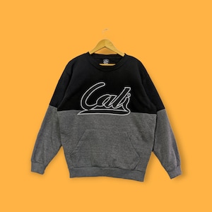 May include: A black and gray sweatshirt with the word "Cali" in white script across the chest. The sweatshirt has a kangaroo pocket and is hanging on a wooden hanger against an orange background.