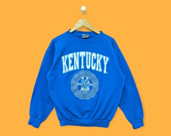 May include: A blue sweatshirt with the text "KENTUCKY" in white on the front. The sweatshirt also features a circular logo with the text "UNIVERSITY OF KENTUCKY" and a graphic of two people shaking hands.