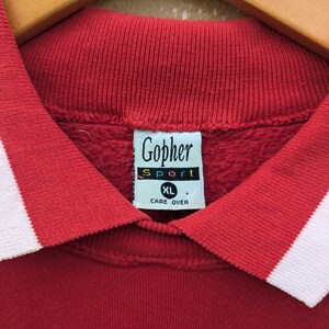 May include: Red collared sweatshirt with white trim. The sweatshirt has a Gopher Sport logo with the text "Care Over" and a size tag that says "XL".