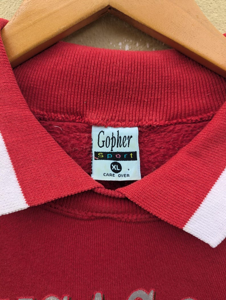 May include: Red collared sweatshirt with white trim. The sweatshirt has a Gopher Sport logo with the text "Care Over" and a size tag that says "XL".