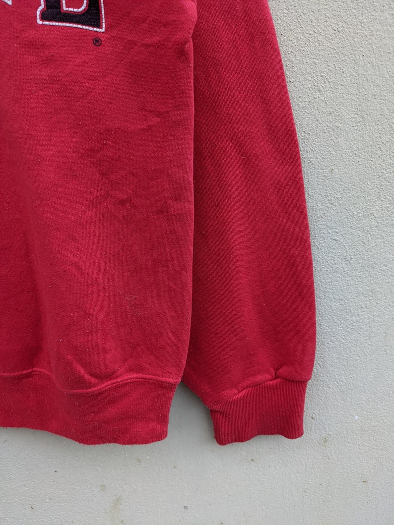 May include: Red sweatshirt with a black and white embroidered logo on the front. The sweatshirt has a ribbed cuff and hem.