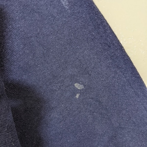 May include: Close-up of a navy blue fabric with visible texture. The fabric has several small, light-colored spots. The image highlights the material's weave and color variations.