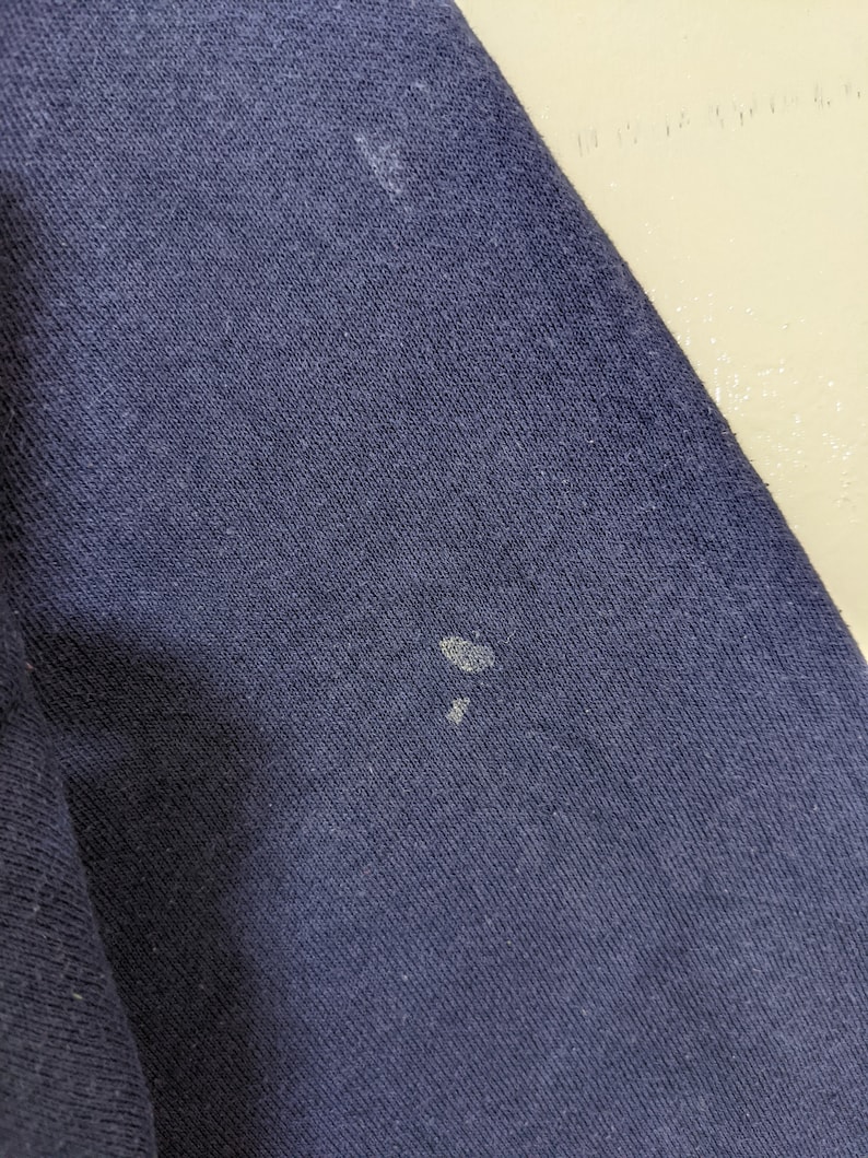 May include: Close-up of a navy blue fabric with visible texture. The fabric has several small, light-colored spots. The image highlights the material's weave and color variations.