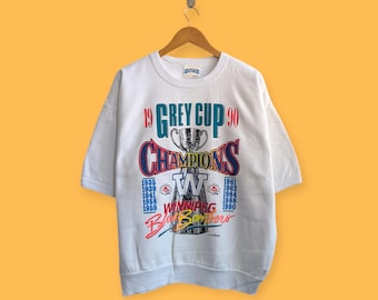 May include: White short-sleeved sweatshirt with a graphic design. The design features the text "19 GREY CUP 90 CHAMPIONS" in red, blue, and white. Below the text is a list of years and the words "WINNIPEG Blue Bombers".