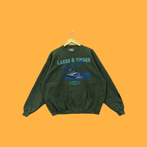 Puede incluir: Una sudadera de color verde oscuro con el texto "LAKES & TIMBER" y un gráfico de un pato en vuelo.