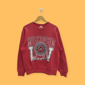 May include: Red sweatshirt with a vintage Wisconsin Badgers logo. The logo features a circular design with the words "University of Wisconsin" and "Numen Lumen" around a stylized eye.