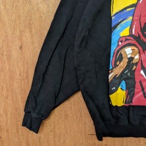 May include: Black sweatshirt with a colorful graphic print of a person wearing a red shirt and a yellow hat. The graphic is in a cartoon style.