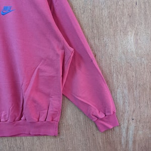 May include: A pink sweatshirt with a blue Nike logo on the front. The sweatshirt has a ribbed collar and cuffs.