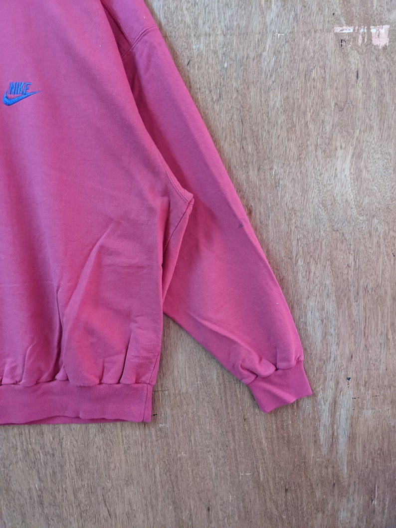May include: A pink sweatshirt with a blue Nike logo on the front. The sweatshirt has a ribbed collar and cuffs.