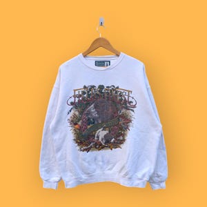 May include: White sweatshirt with a hunting-themed graphic. The graphic features a brown bear, a hunting dog, and foliage. The sweatshirt is hanging on a wooden hanger against a yellow background. The text on the graphic is partially visible.