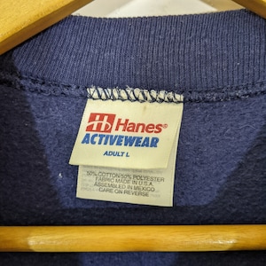 May include: Close-up of a navy blue sweatshirt collar with a Hanes Activewear tag. The tag indicates the size is Adult L and the fabric is 50% cotton and 50% polyester. The garment was assembled in Mexico.