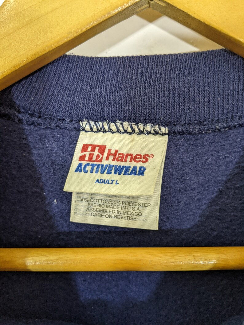 May include: Close-up of a navy blue sweatshirt collar with a Hanes Activewear tag. The tag indicates the size is Adult L and the fabric is 50% cotton and 50% polyester. The garment was assembled in Mexico.