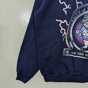 May include: Navy blue sweatshirt featuring a circular emblem with the text "UNIVERSITY OF NORTH CAROLINA" and a cartoon mascot. White lightning bolt graphics are on the upper left. The text "GO TAR HEELS" is below the emblem.