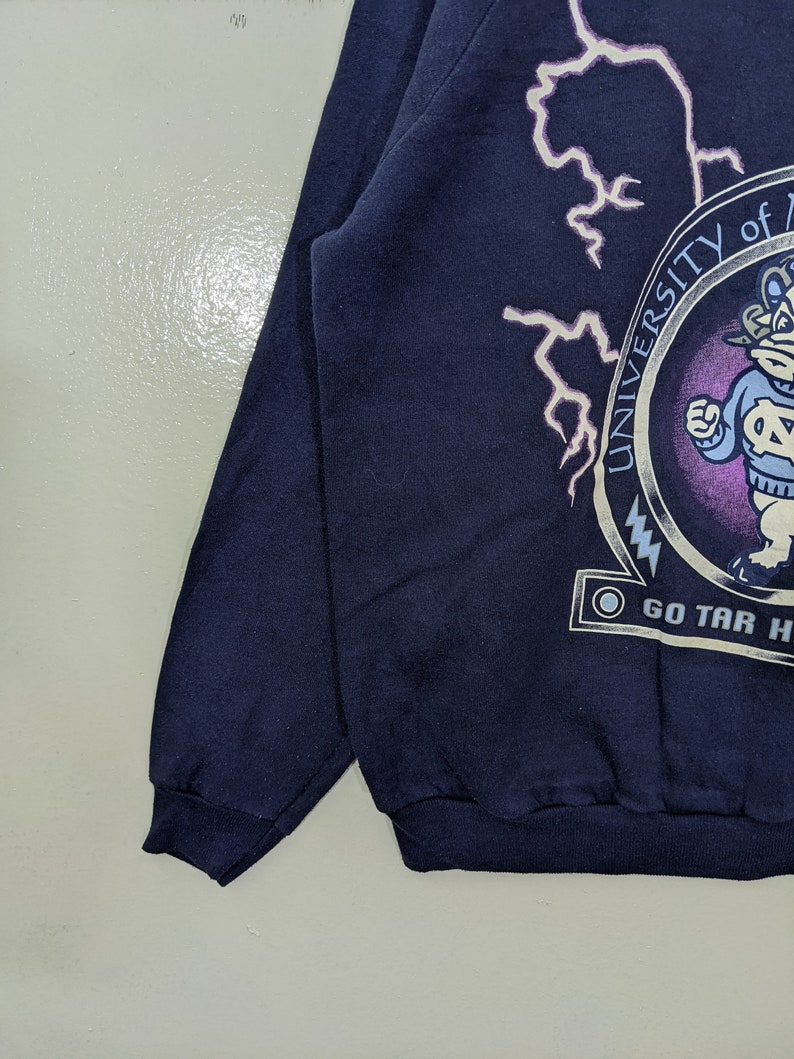 May include: Navy blue sweatshirt featuring a circular emblem with the text "UNIVERSITY OF NORTH CAROLINA" and a cartoon mascot. White lightning bolt graphics are on the upper left. The text "GO TAR HEELS" is below the emblem.