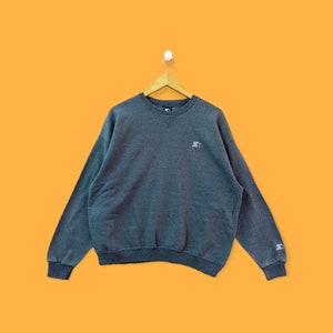 Puede incluir: Una sudadera gris con un pequeño logotipo en el pecho izquierdo. La sudadera tiene cuello redondo y mangas largas.