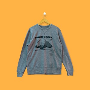 Puede incluir: Sudadera gris con un estampado negro de un edificio y el texto "Knob Creek Bourbon Whiskey" y "There are no shortcuts out of Clermont, KY".