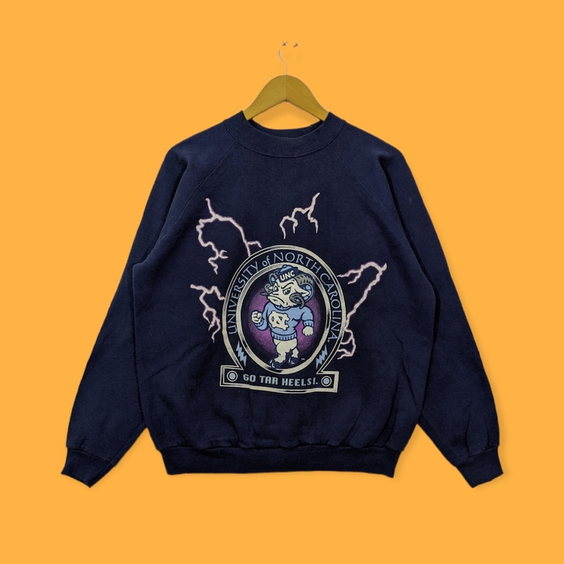 May include: Navy blue sweatshirt featuring a graphic of the University of North Carolina mascot inside a circle, with the text "UNIVERSITY OF NORTH CAROLINA" and "GO TAR HEELS!". The design includes lightning bolts and a map outline.