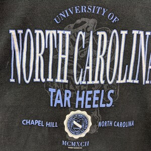 Puede incluir: Sudadera negra con el texto "UNIVERSITY OF NORTH CAROLINA TAR HEELS" en blanco y azul claro. El dise&ntilde;o incluye un sello con el texto "CHAPEL HILL NORTH CAROLINA" y el n&uacute;mero romano "MCMXCII".