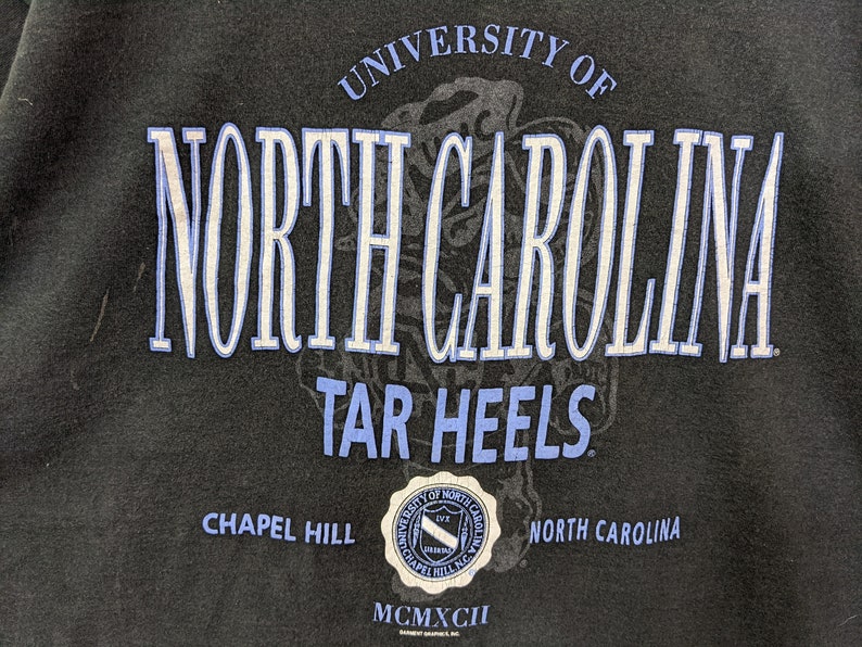 Puede incluir: Sudadera negra con el texto "UNIVERSITY OF NORTH CAROLINA TAR HEELS" en blanco y azul claro. El dise&ntilde;o incluye un sello con el texto "CHAPEL HILL NORTH CAROLINA" y el n&uacute;mero romano "MCMXCII".