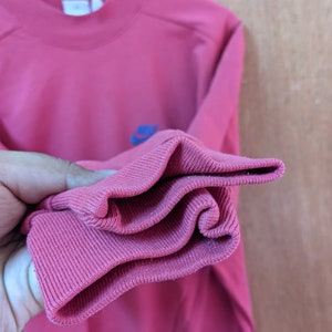 May include: A pink sweatshirt with a blue and white logo on the front. The sweatshirt is folded up to show the ribbed cuffs.