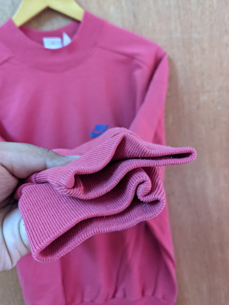 May include: A pink sweatshirt with a blue and white logo on the front. The sweatshirt is folded up to show the ribbed cuffs.