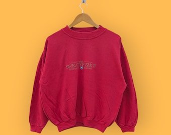 May include: Red sweatshirt with a white embroidered logo that reads "The Sweater Shop" with a small blue and white cross.