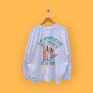 Op de afbeelding: Wit sweatshirt met een afbeelding van surfplanken en de tekst "Le Bonheur de Skier Club Med".
