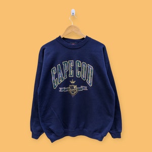 May include: A navy blue sweatshirt with a vintage-style graphic of "CAPE COD" in a green, yellow, and purple font. The graphic also includes a crest with a crown and a banner.