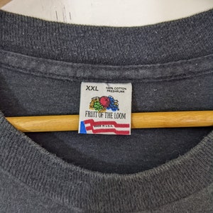 Puede incluir: Primer plano de una camiseta Fruit of the Loom XXL gris oscuro. La etiqueta dice "100% algod&oacute;n pre-encogido" y "Made in U.S.A.". La camiseta est&aacute; colgada en una percha de madera.