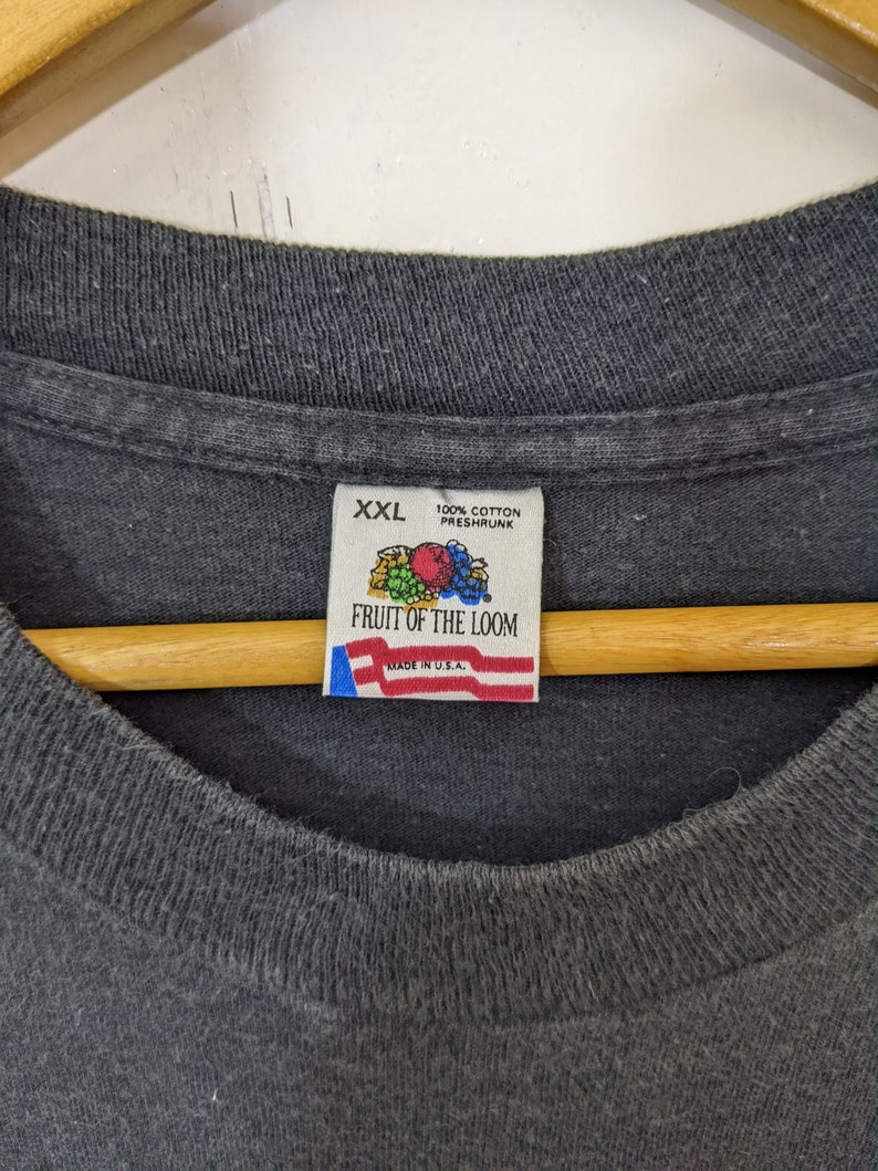 Puede incluir: Primer plano de una camiseta Fruit of the Loom XXL gris oscuro. La etiqueta dice "100% algod&oacute;n pre-encogido" y "Made in U.S.A.". La camiseta est&aacute; colgada en una percha de madera.