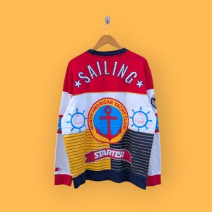 以下が含まれることがあります： 上部に「SAILING」の文字が入った、赤、白、青のヴィンテージスウェットシャツ。アンカーと「North American Yacht Club」と「Starter」の文字が入った、航海をテーマにしたデザインです。