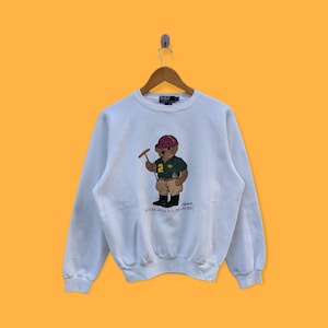 May include: White crewneck sweatshirt with a brown bear wearing a red hat and holding a polo mallet. The bear is wearing a green polo shirt with the number 2 on it. The text "Polo Bear by Ralph Lauren" is printed below the bear.