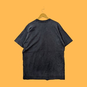 Puede incluir: Una camiseta negra colgada de una percha de madera sobre un fondo naranja. La camiseta es de color carb&oacute;n oscuro y parece estar hecha de un material suave y desgastado. La camiseta es de cuello redondo.