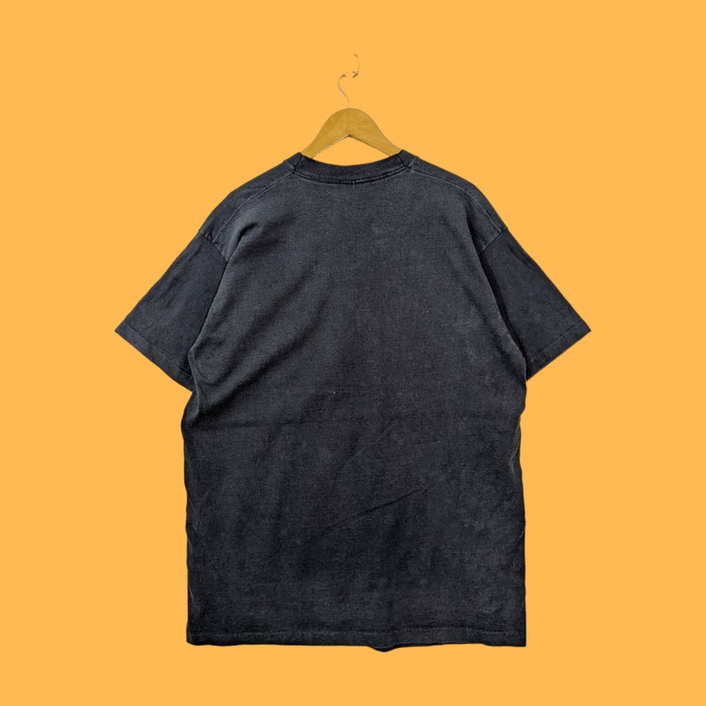 Puede incluir: Una camiseta negra colgada de una percha de madera sobre un fondo naranja. La camiseta es de color carb&oacute;n oscuro y parece estar hecha de un material suave y desgastado. La camiseta es de cuello redondo.