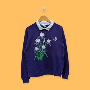 Puede incluir: Una sudadera morada con cuello blanco y un diseño floral con iris blancos y morados y una mariposa.