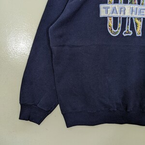 Peut inclure: Un sweat-shirt bleu marine avec un patch blanc qui dit "TAR HEELS" et un motif paisley bleu et blanc qui dit "UNC".