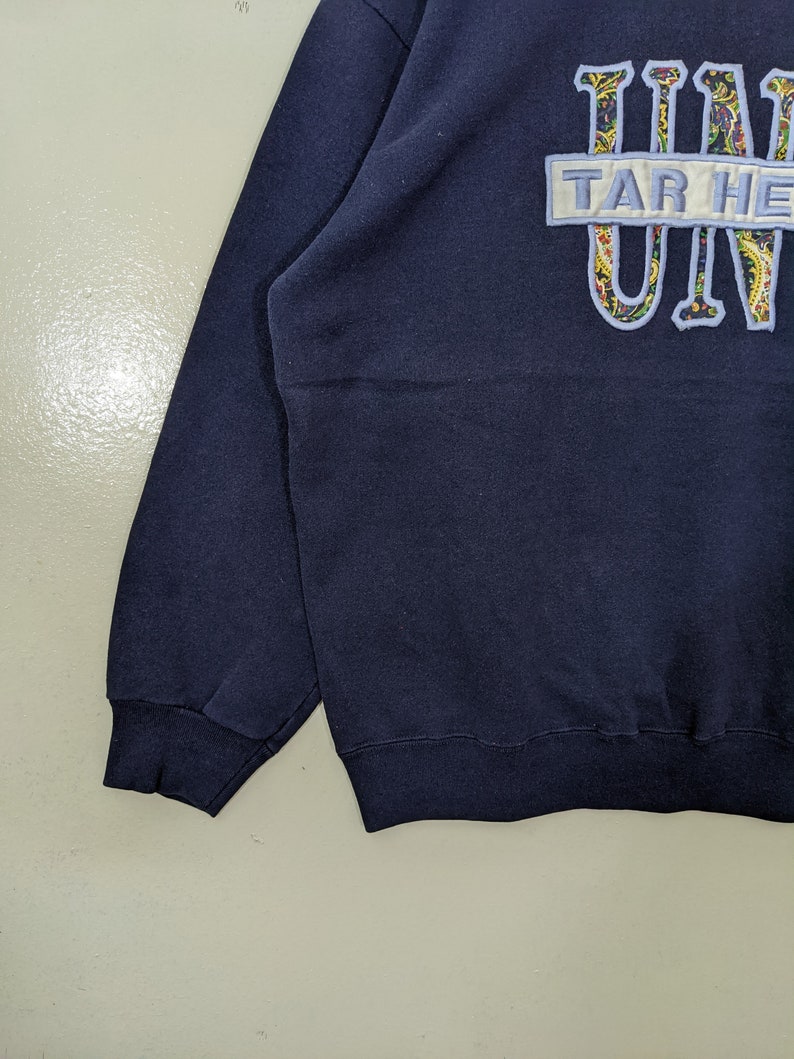 Peut inclure: Un sweat-shirt bleu marine avec un patch blanc qui dit "TAR HEELS" et un motif paisley bleu et blanc qui dit "UNC".