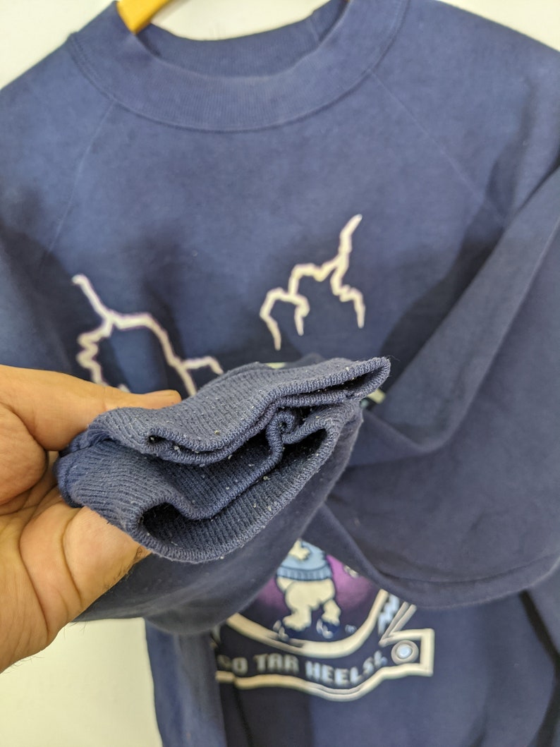 May include: Navy blue sweatshirt with white lightning bolt graphics and a collegiate emblem. The emblem features a cartoon bear wearing a sweater and the text "GO TAR HEELS". The sweatshirt has a crew neck and long sleeves.