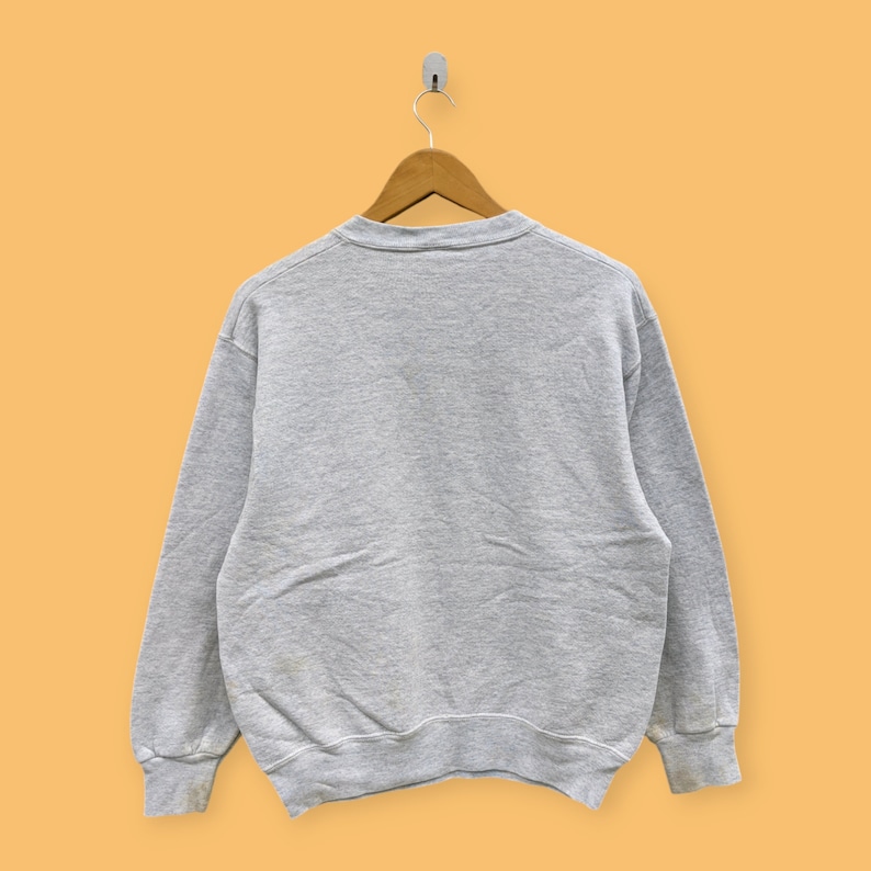 May include: A gray sweatshirt with long sleeves and a crew neck. The sweatshirt is hanging on a wooden hanger against a yellow background.