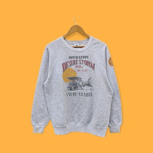 Peut inclure: Un sweat-shirt gris avec l'inscription "OPERATION DESERT STORM" en rouge et blanc. En dessous, une illustration de véhicules militaires et les mots "SAUDI ARABIA". Le sweat-shirt a un col rond et des manches longues.