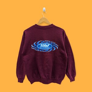 Peut inclure: Un sweat-shirt bordeaux à col ras du cou, suspendu à un cintre en bois sur un fond orange. Le dos du sweat-shirt présente un motif bleu et blanc avec le texte "World Jungle".