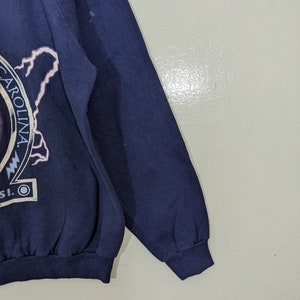 May include: Navy blue sweatshirt with a graphic featuring the word "CAROLINA" in a circular design, a lightning bolt, and other text. The sweatshirt has a ribbed cuff and hem.