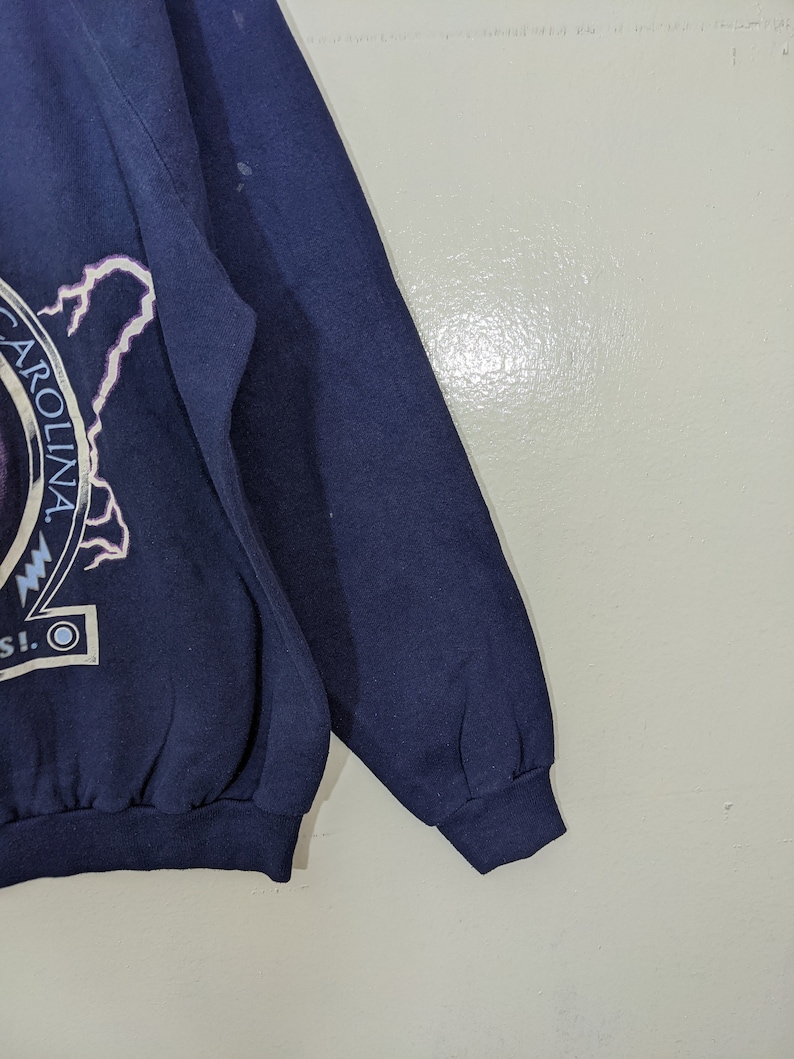 May include: Navy blue sweatshirt with a graphic featuring the word "CAROLINA" in a circular design, a lightning bolt, and other text. The sweatshirt has a ribbed cuff and hem.