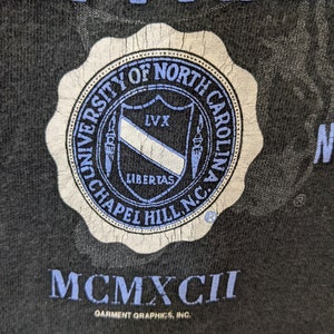 Puede incluir: Primer plano de una camiseta negra con un sello desgastado de la Universidad de Carolina del Norte en Chapel Hill en azul y blanco. El texto "MCMXCII" est&aacute; debajo del sello, con "Garment Graphics, Inc." impreso debajo.